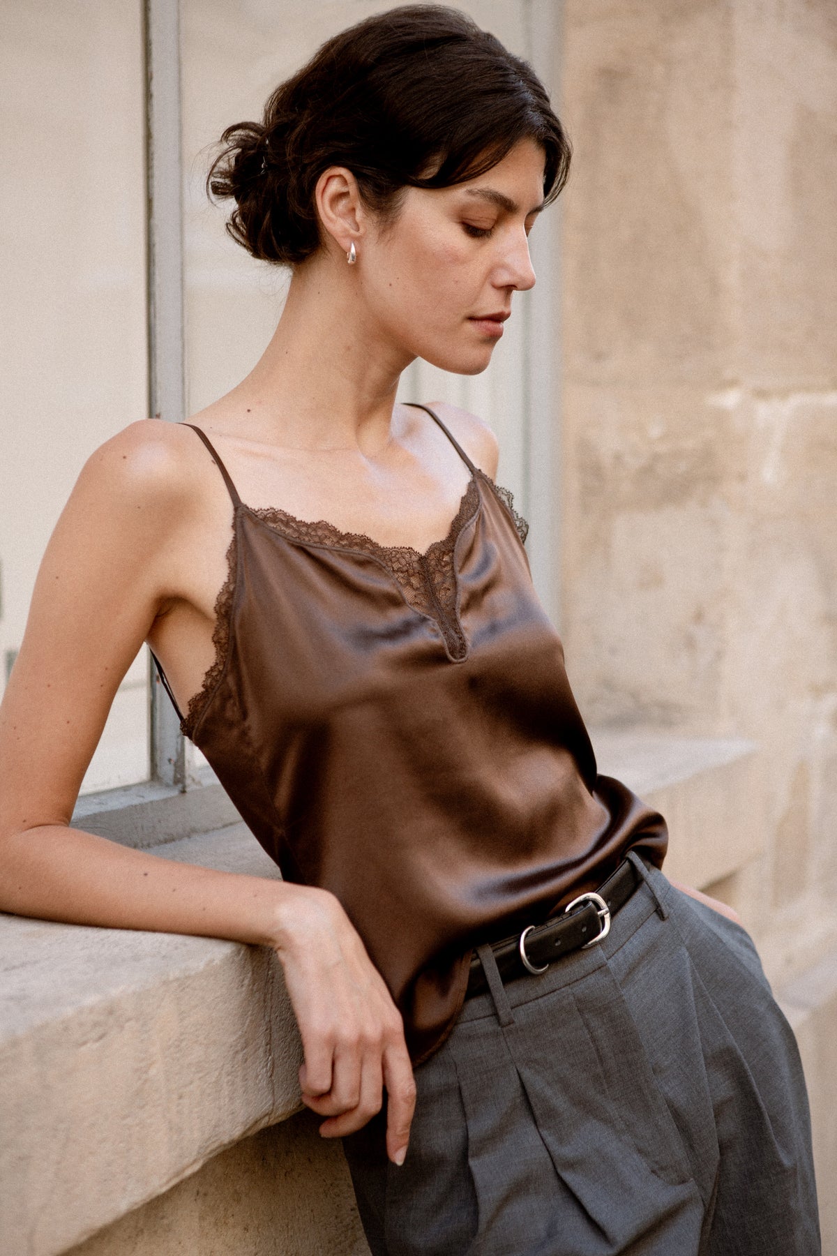 A woman wearing a brown silk camisole and dark grey wide-leg trousers, leaning against a stone wall.