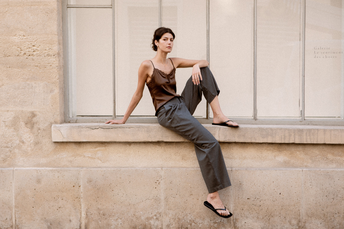 A woman sits on a stone window ledge of a beige building, wearing a brown silk camisole, dark grey wide-leg trousers, with black sandals.
