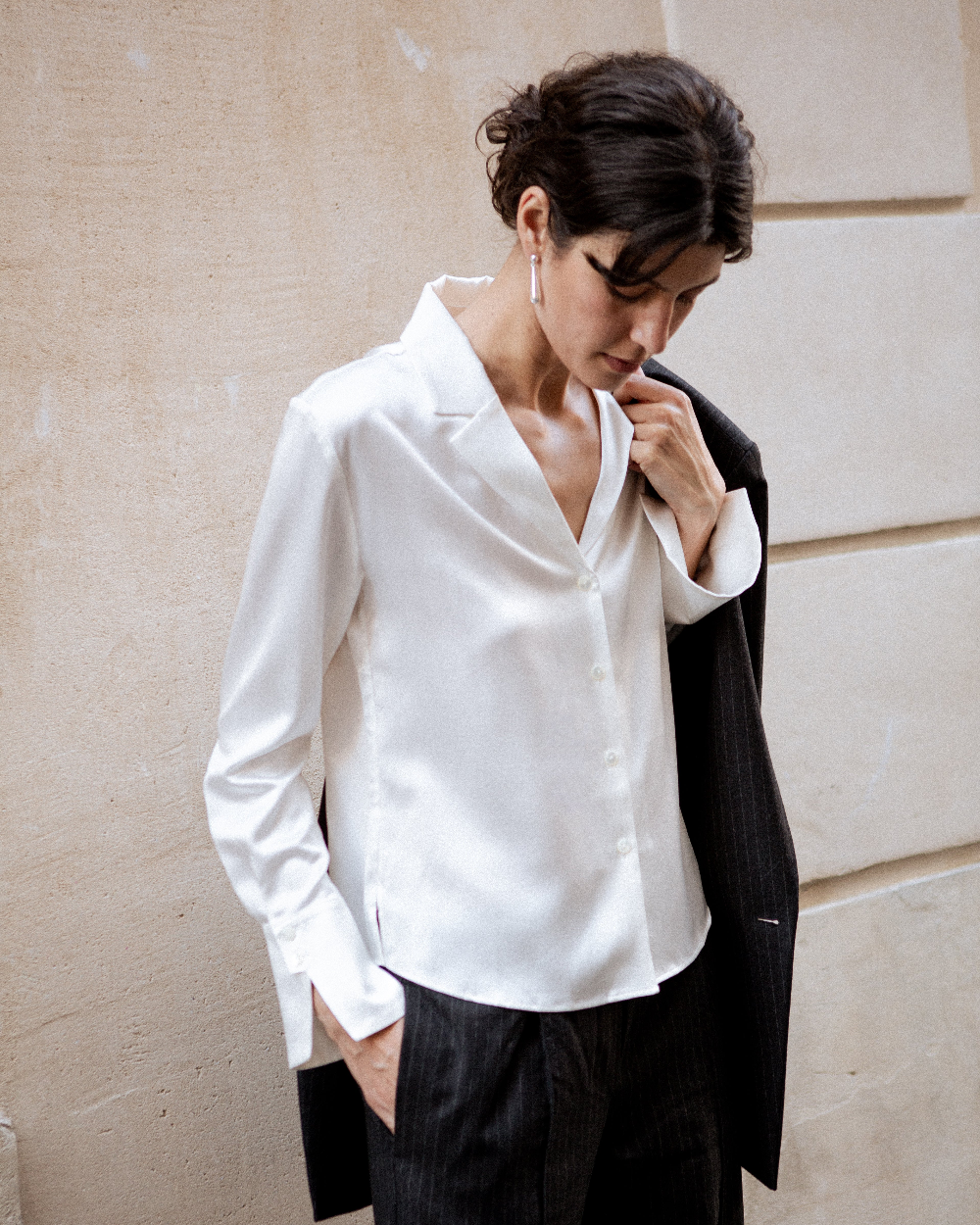 A woman wearing a white long-sleeve button-up shirt and dark pinstriped trousers, holding a black blazer against her shoulder, standing against a beige stone wall.