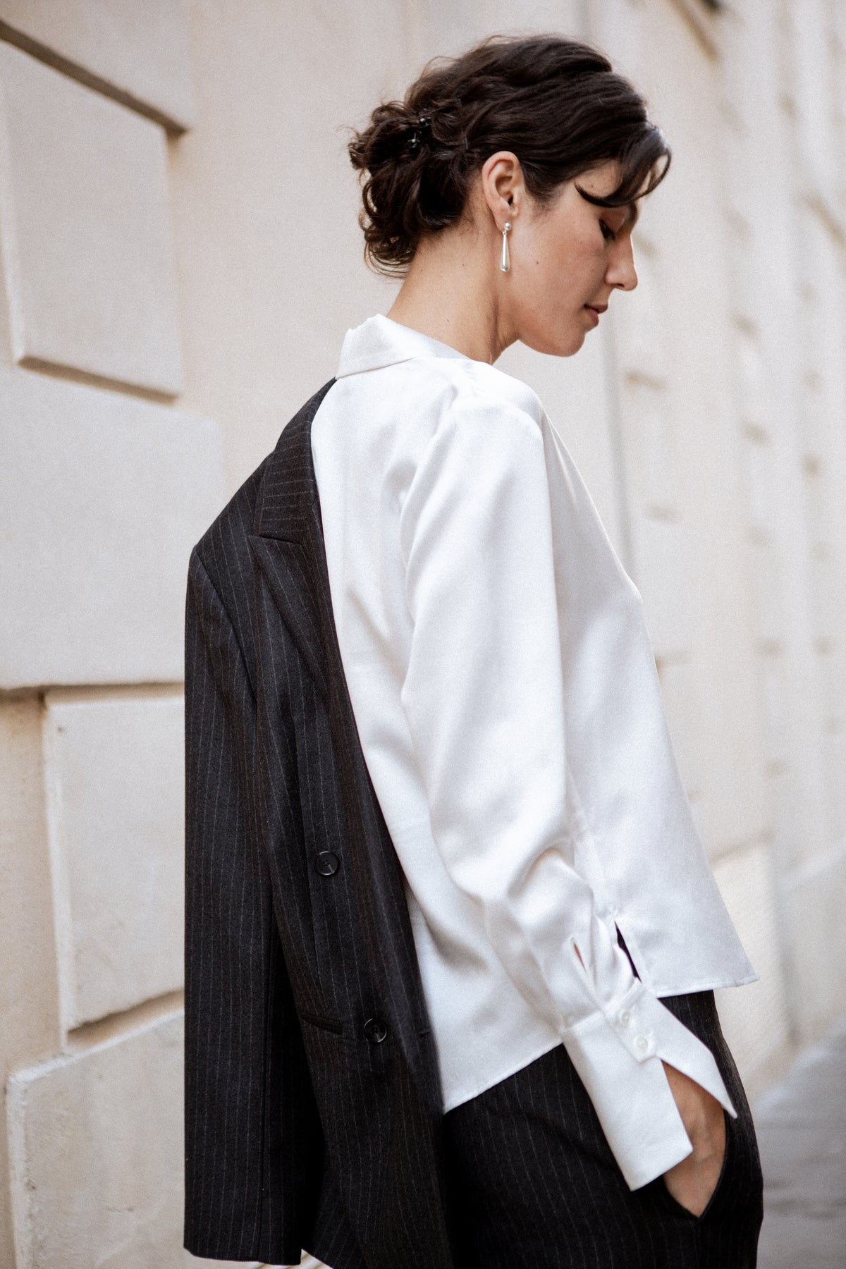 A woman wearing a white long-sleeve silk shirt blouse with a black pinstripe blazer draped over her shoulders, standing against a light beige stone wall.