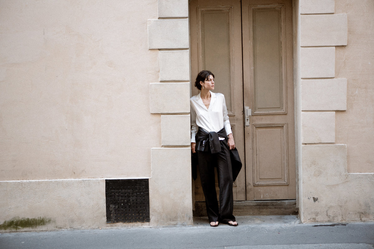 A woman stands in a doorway wearing a white button-up silk shirt, dark wide-leg trousers, and black sandals, with a dark jacket tied around her waist.